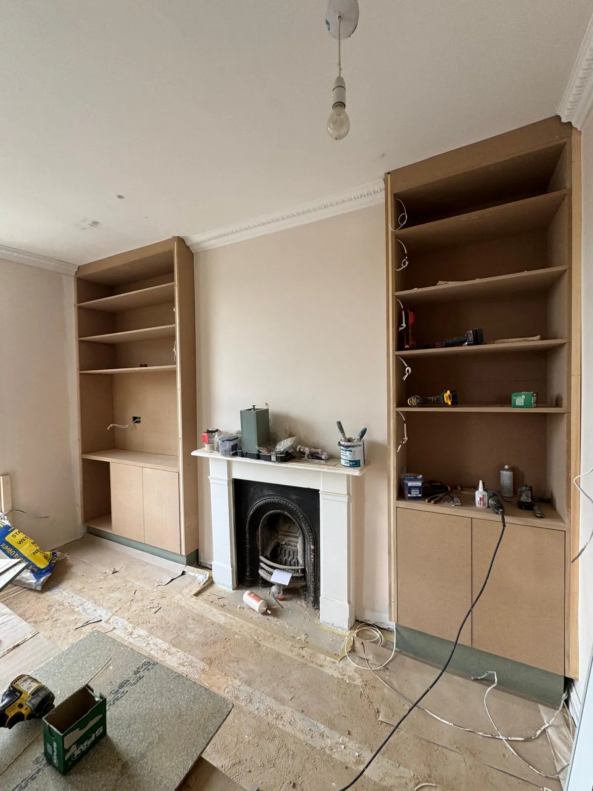 Living room under construction with newly installed MDF alcove shelving units flanking a Victorian cast-iron fireplace