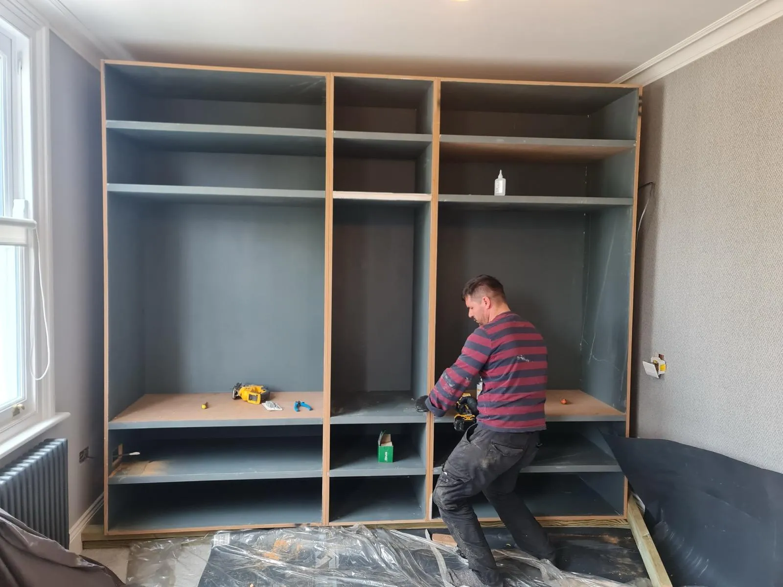 Carpenter installing large grey-blue fitted wardrobe carcass with open shelving and hanging rail in a wallpapered bedroom