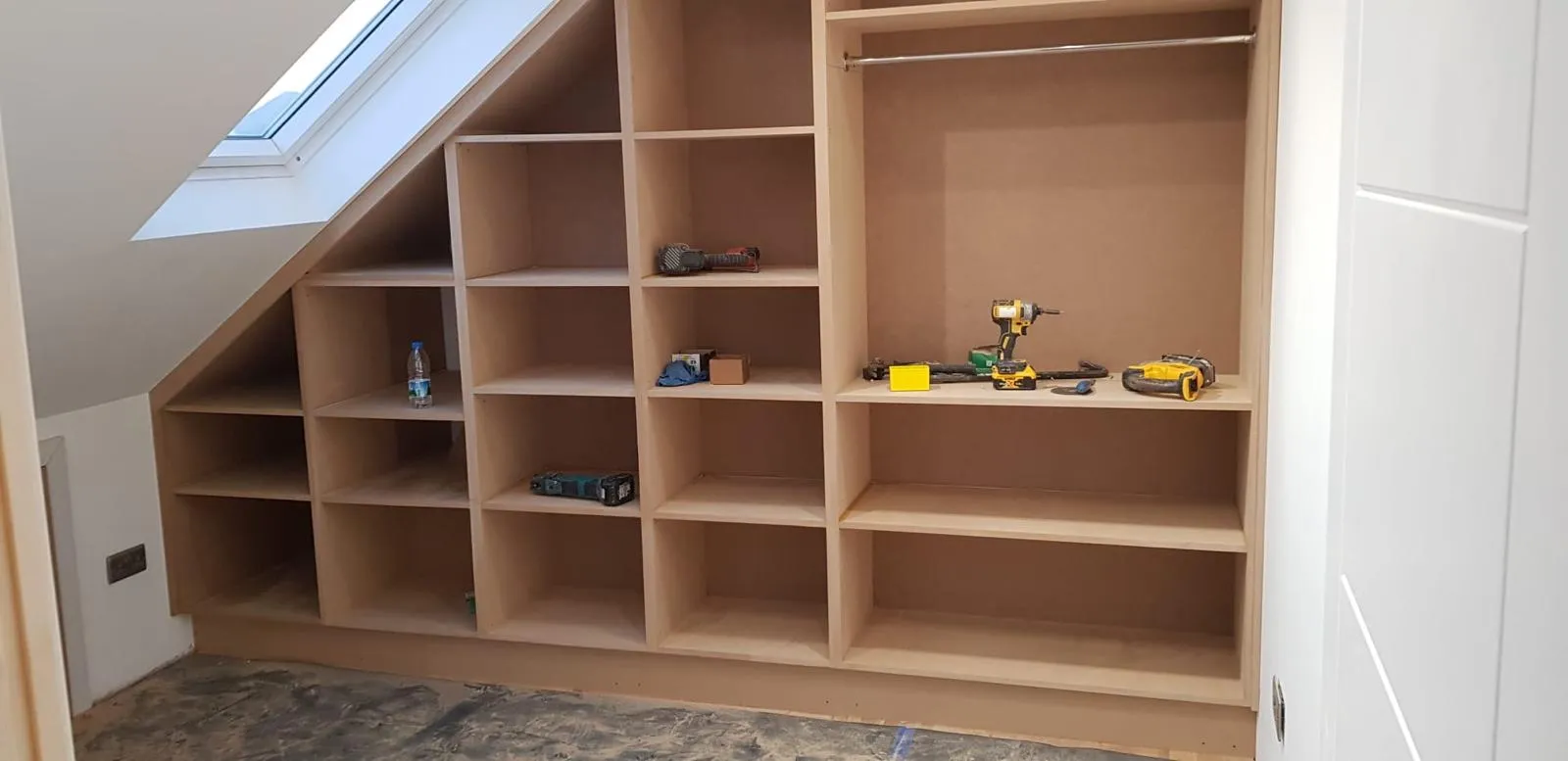 Attic room under fit-out with bespoke stepped MDF shelving and wardrobe built to follow the sloping roofline under Velux skylights