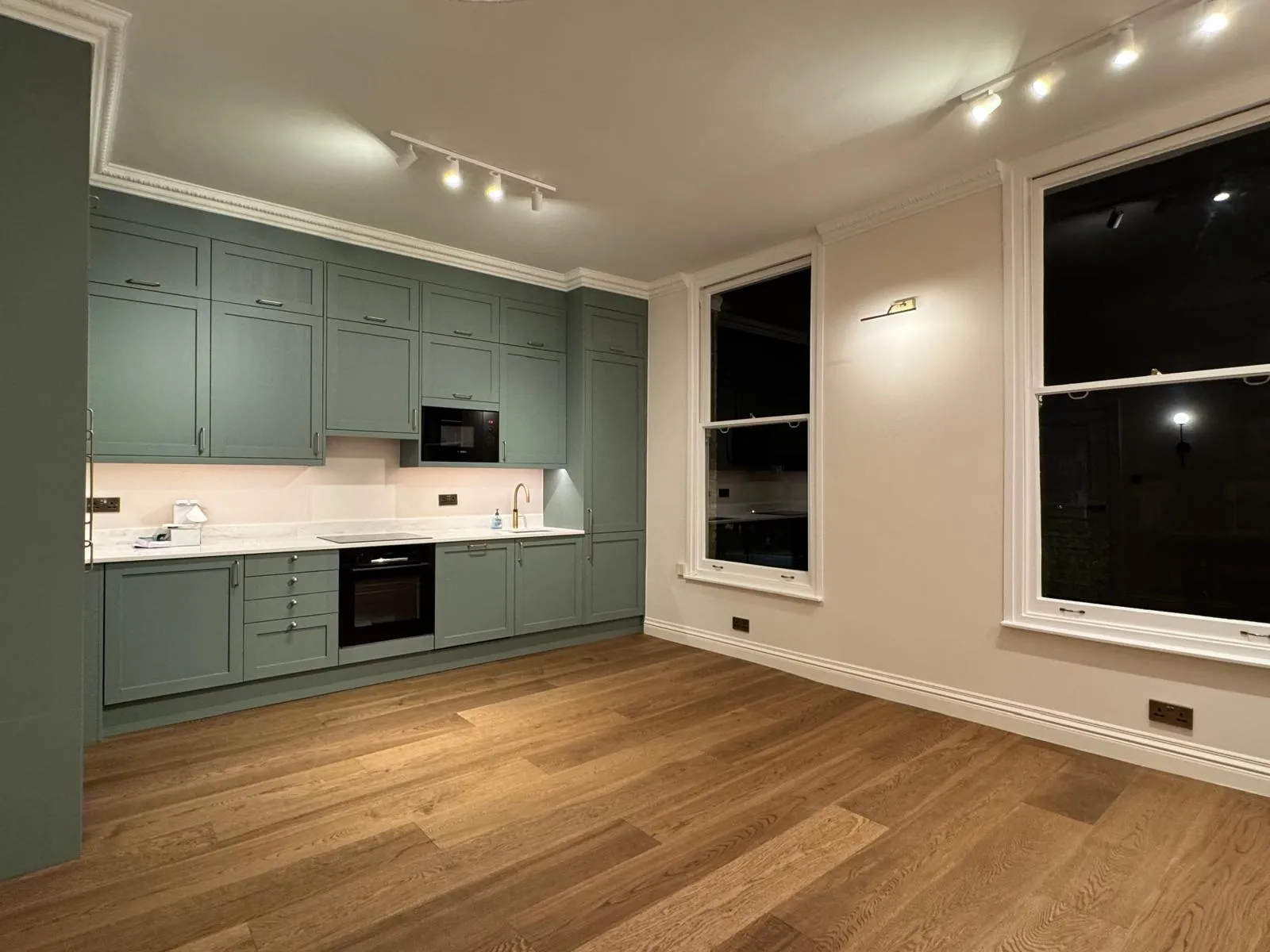 Finished studio kitchen with teal-green shaker cabinetry, stone worktop, integrated appliances, brass tap and oak plank floor