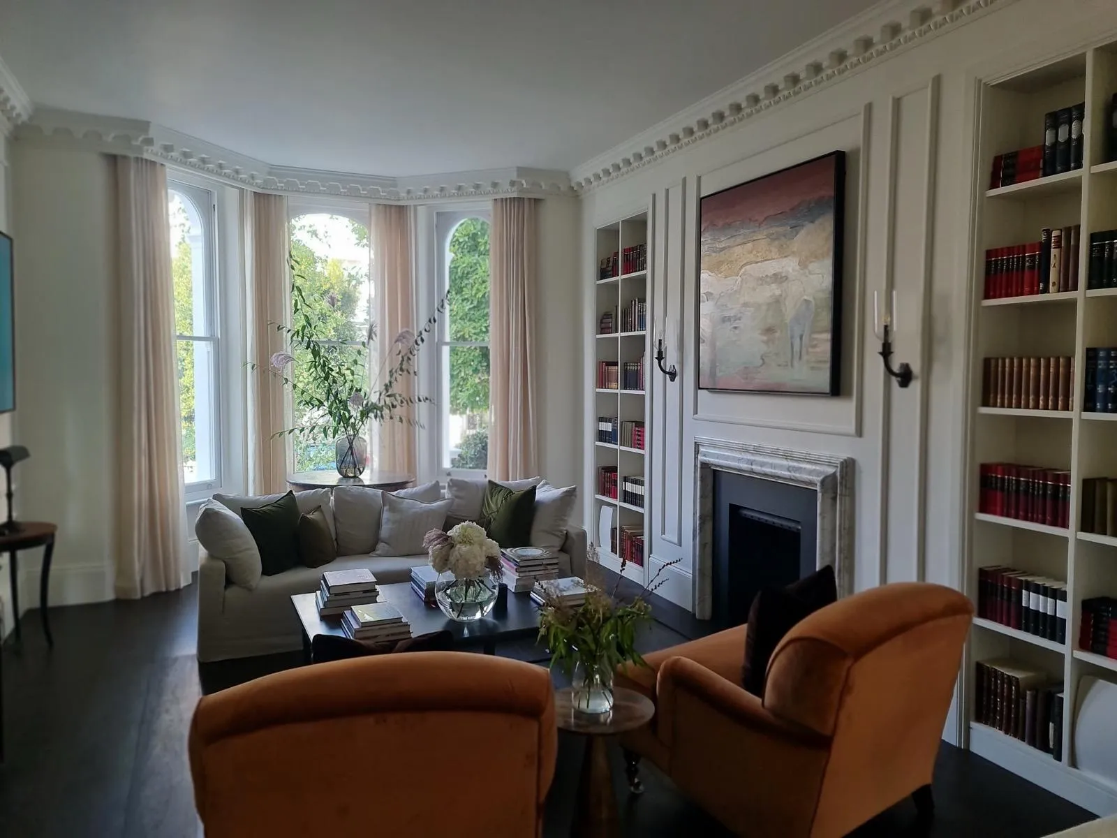 Finished drawing room with bay window, linen sofa, rust velvet armchairs, marble fireplace and book-lined alcove shelves