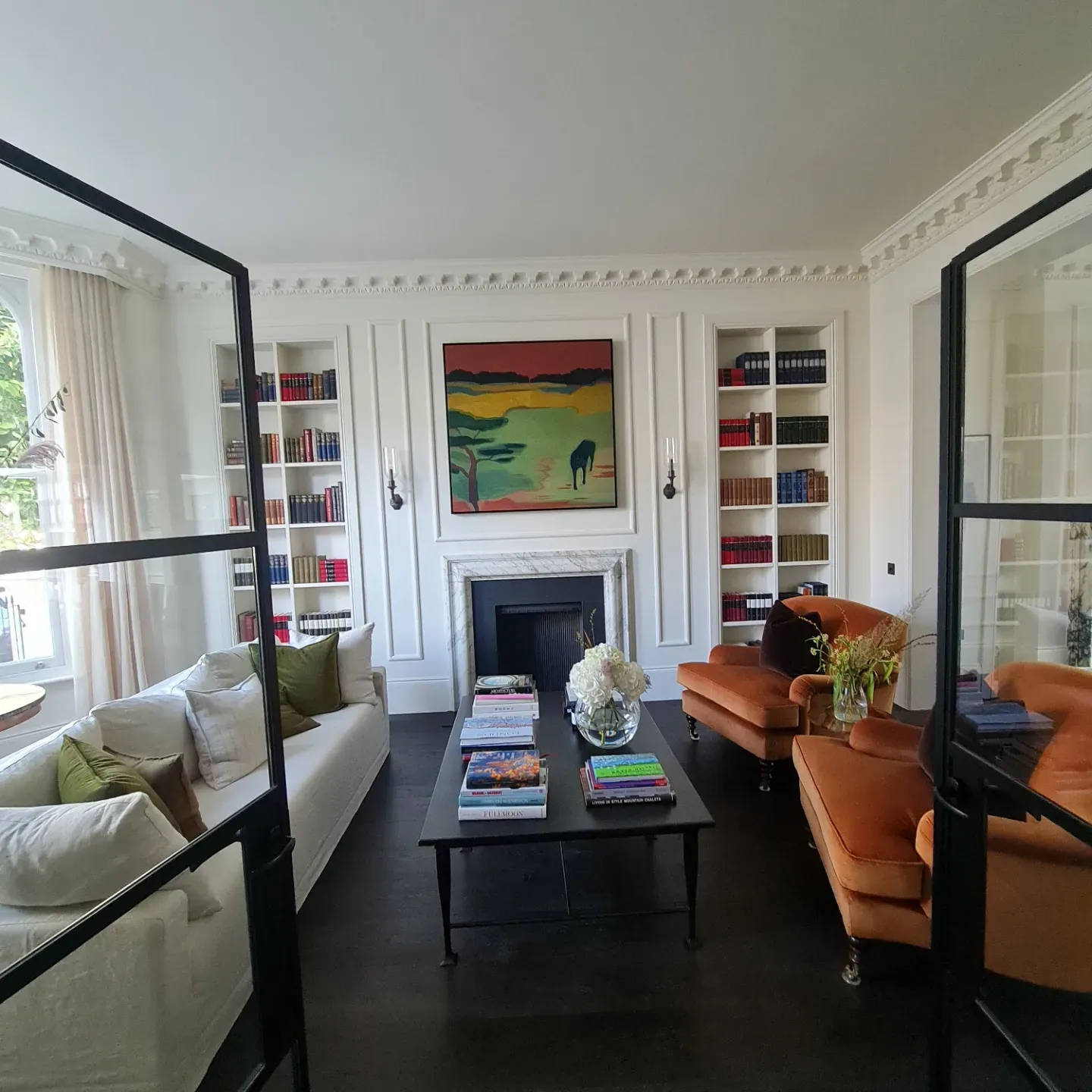 Finished drawing room with white marble fireplace, built-in bookshelves, rust velvet armchairs and dark oak floor viewed through crittall doors