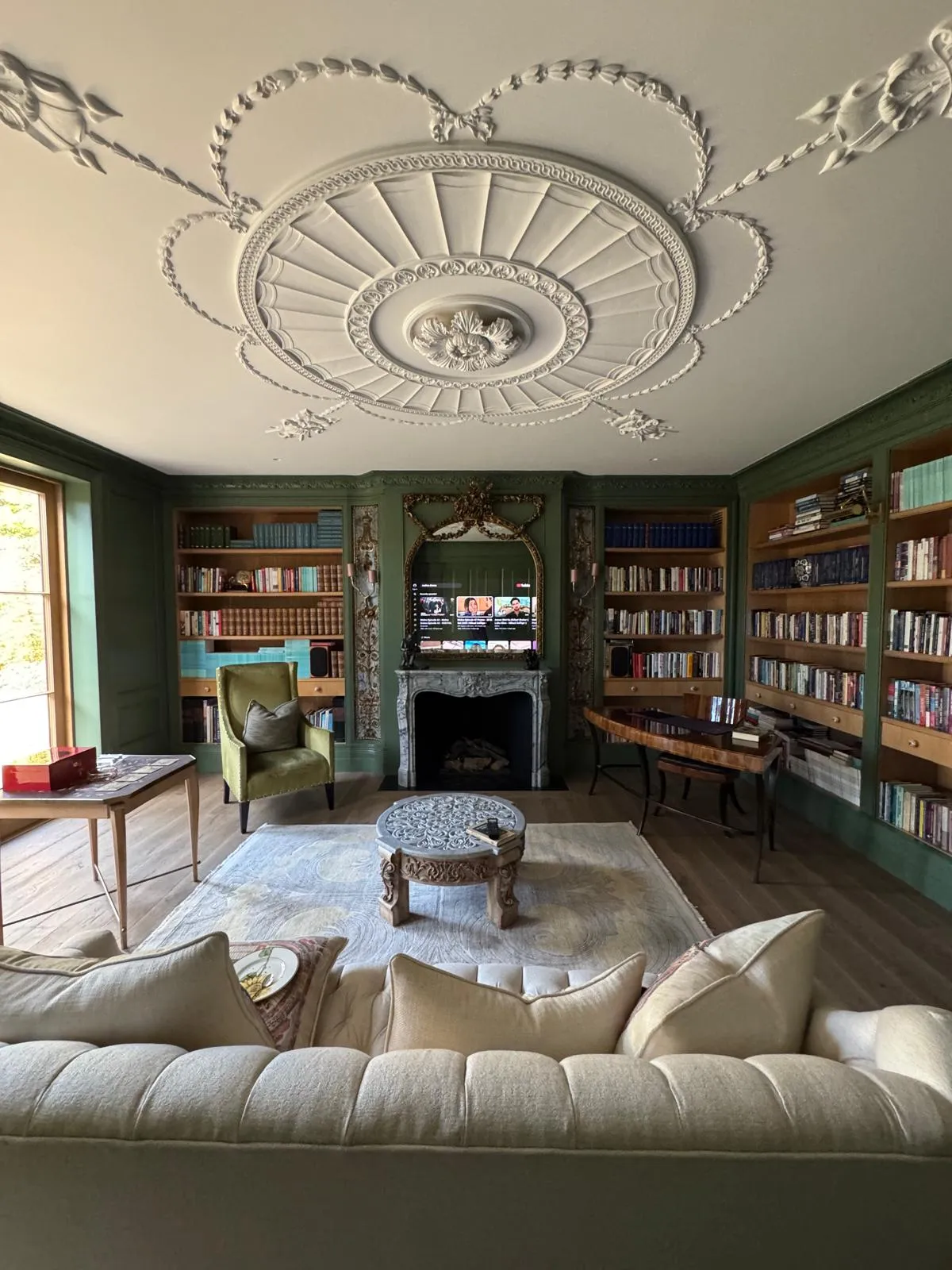 Finished library with green panelled walls, ornate plaster ceiling rose, floor-to-ceiling bookshelves, marble fireplace and linen sofa