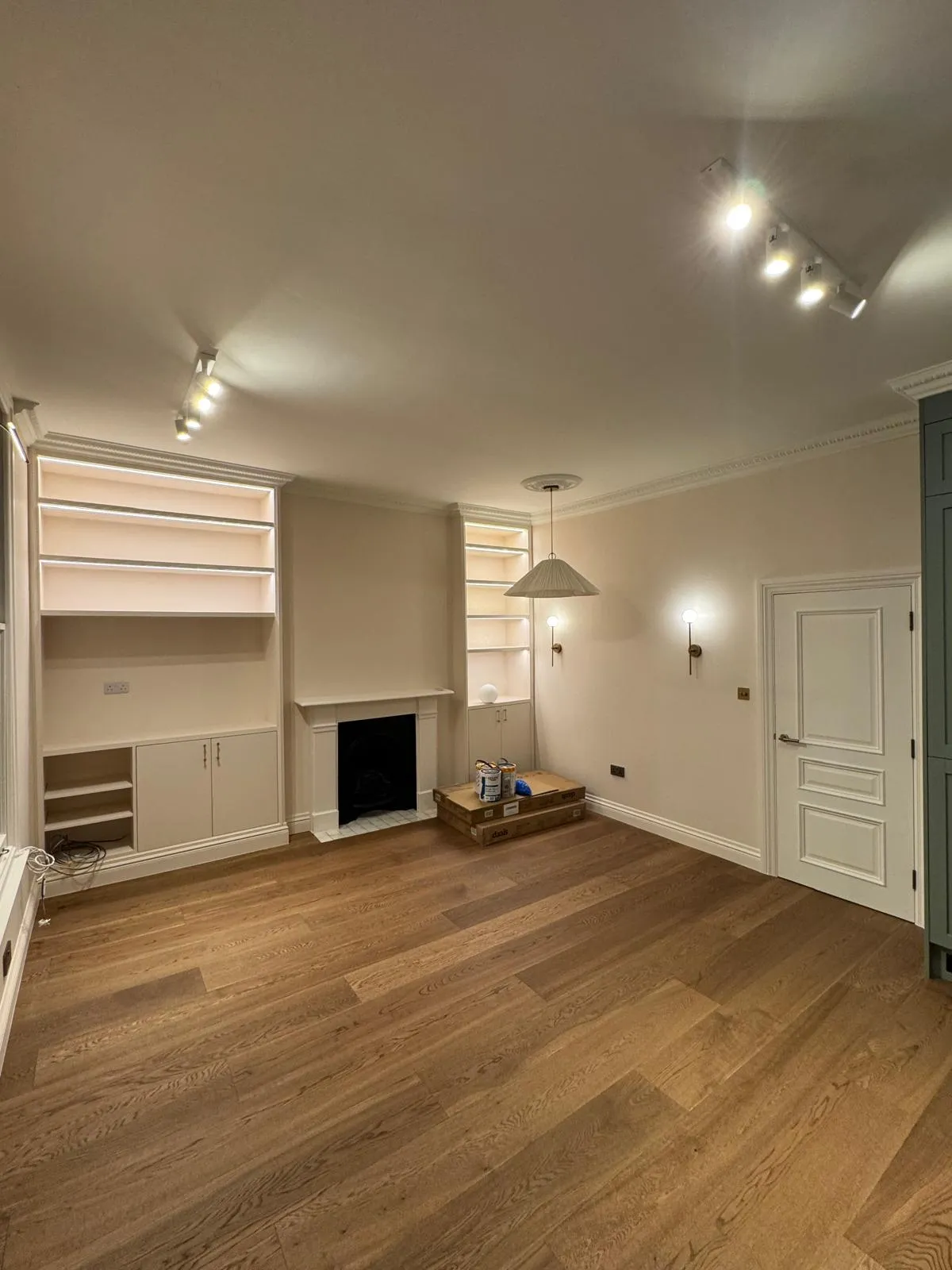 Nearly finished reception room with cream alcove shelving, black Victorian fireplace, oak plank flooring and LED track lighting