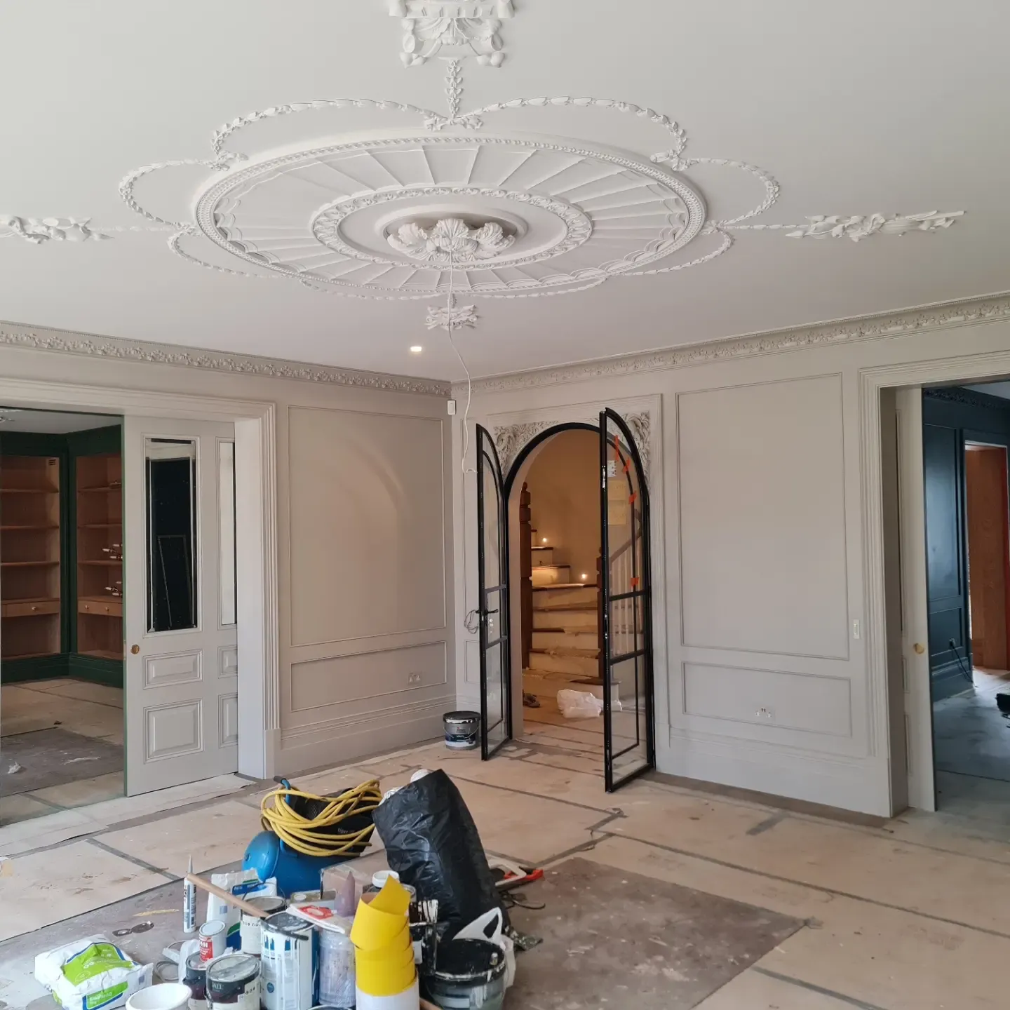 Reception room under renovation featuring elaborate plaster ceiling rose, panelled walls and arched black-framed glazed doors